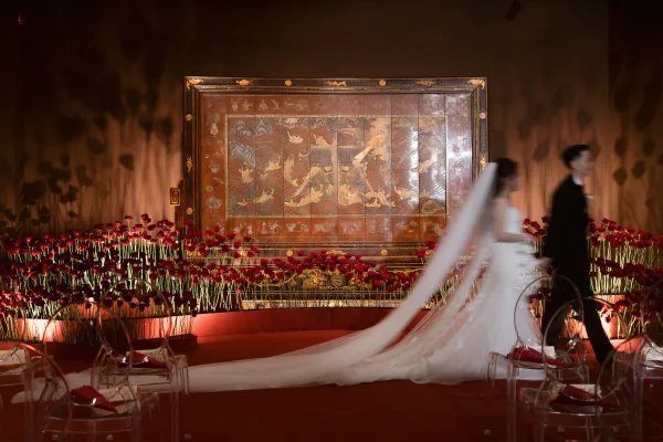 Ceremony stage decor with red rose ceremony backdrop, warm uplighting and clear acrylic chairs as bride and groom pass the folding screen backdrop