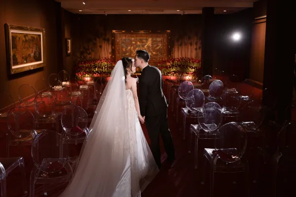Wedding couple portrait of groom kissing bride’s forehead as they hold hands, her veil train flowing down a red aisle runner in a moody indoor ceremony room