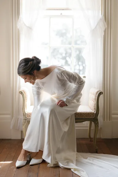 Bridal getting ready bride putting on shoes in a satin long-sleeve wedding dress on a vintage bench by a large window with sheer curtains