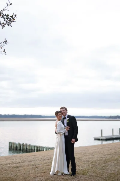 Couple portrait of bride in a long sleeve wedding dress and groom in tuxedo with boutonniere embracing on a waterfront dock under cloudy sky