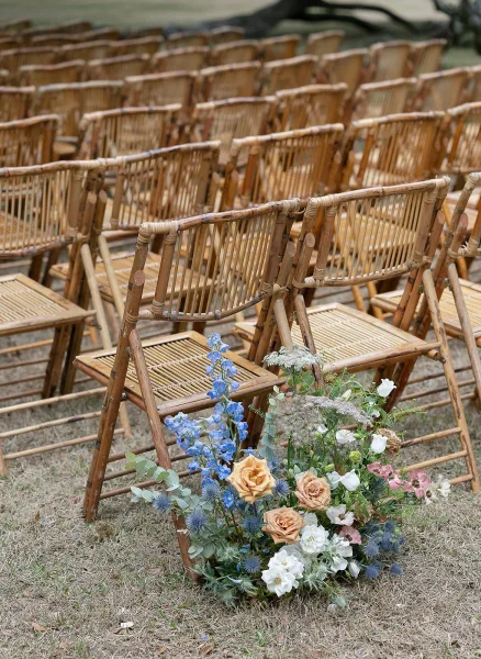Ceremony seating with outdoor ceremony chairs in neat rows on a grass lawn, lined with a rose and blue delphinium floral arrangement with eucalyptus