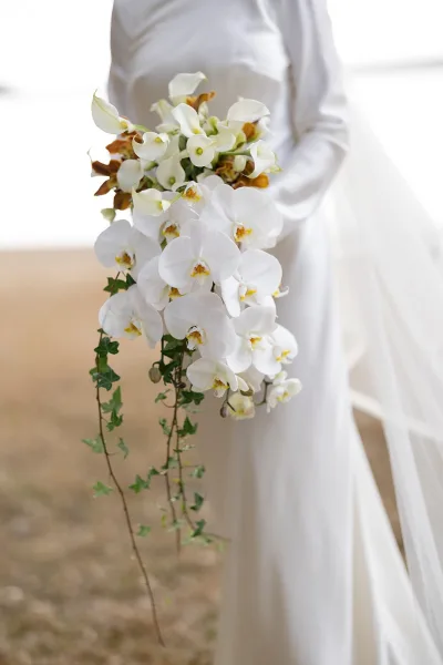 Bridal bouquet of white orchid bouquet and calla lilies cascading with ivy vines in the bride’s hands, veil and bright sky behind