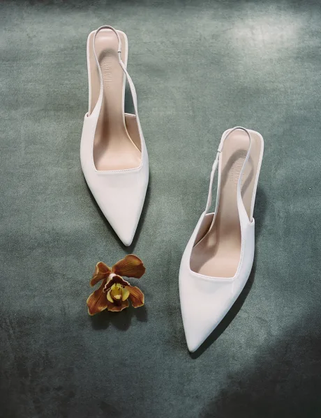 Wedding shoes, white bridal heels in a flat lay with an orchid flower accent on a dark textured surface