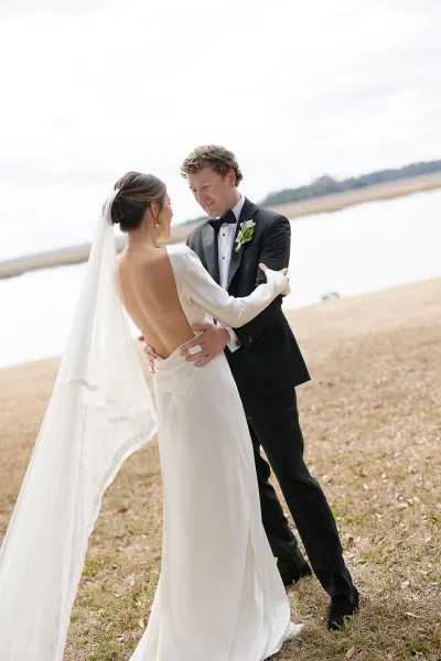 Couple portrait of bride and groom embrace by a lakeshore, her cathedral veil blowing in wind as he holds her waist under cloudy sky