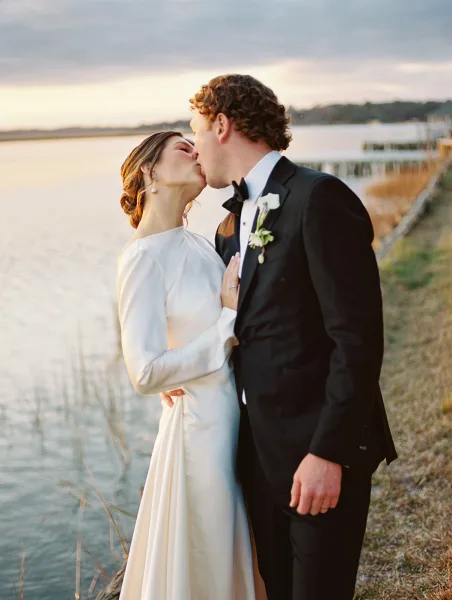 Wedding kiss as bride in a long sleeve dress and groom in a tuxedo embrace on a dock by the waterfront under a cloudy sky