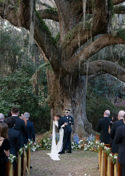 Ceremony kiss between bride and groom at an outdoor wedding ceremony under a large oak tree with hanging moss and a rose petal aisle