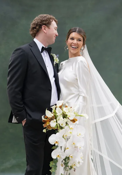 Couple portrait of a bride laughing in a satin wedding dress and veil beside groom in black tuxedo, holding orchid bouquet by a green lake