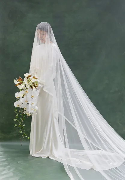 Bridal portrait of a bride in a minimalist wedding dress with a long veil over her face, holding an orchid bouquet on a green studio backdrop