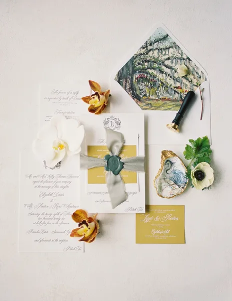 Invitation suite wedding invitation flat lay with envelopes, wax seal, silk ribbon, orchids and greenery, plus an oyster shell ring dish on white surface