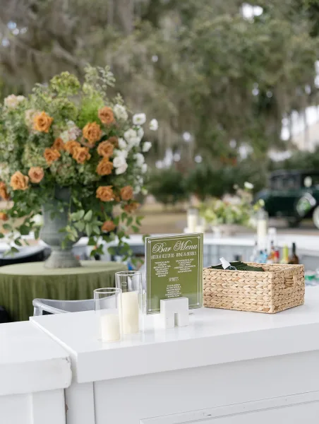 Wedding bar setup with a wedding bar menu sign, floral arrangement, pillar candles and wine bottles on a white counter under mossy trees