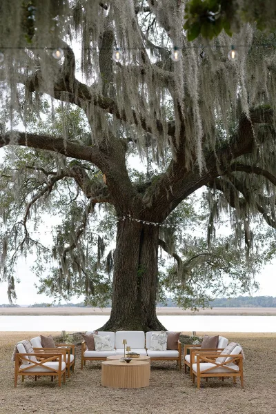 Outdoor lounge setup with a wedding lounge area sofa and chairs, candles and pillows under an oak tree with Spanish moss by the waterfront