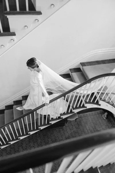 Bridal portrait of a bride on staircase in a long sleeve wedding dress, veil train flowing along carpeted steps by white walls