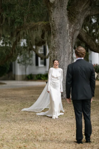 First look moment as groom sees bride in long sleeve wedding dress and cathedral veil beneath oak tree with hanging moss on lawn