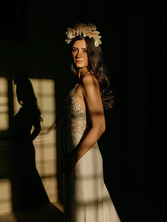 Bridal portrait of a side profile bride in a lace wedding dress with a floral headpiece, lit by window light in a dark room with wall molding