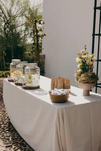 Welcome drink station arranged as an infused water bar with citrus slices, set against a white wall with garden greenery along a stone path.