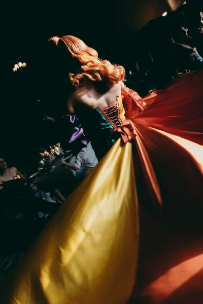 Colorful wedding dress, a rainbow bridal gown with corset lacing, in a dim reception hall among guests and banquet tables with floral centerpieces.