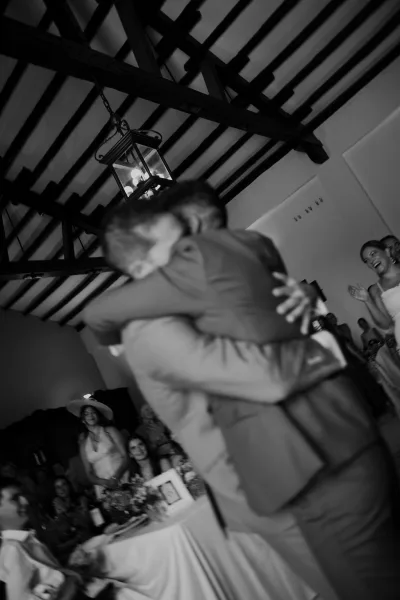 Reception moment capturing a wedding hug moment between friends in suits, beneath an exposed beam ceiling with white walls and seated guests.