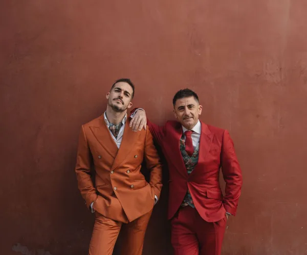Grooms portrait in two grooms style with colorful wedding suits, one wearing a neck scarf, against a terracotta stucco wall