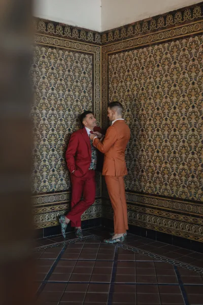Couple portrait of two grooms getting ready, one adjusting his tie, against a patterned tile wall and floor.