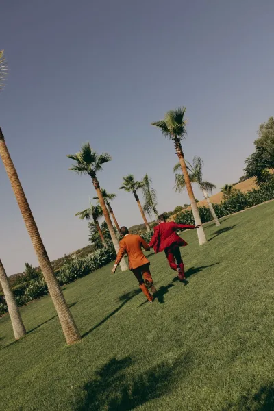Couple portrait of grooms holding hands on a lawn with palm trees and blue sky, their suits adding crisp lines to the joyful scene.
