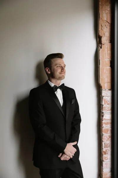 Groom portrait of a black tuxedo groom by a white wall and exposed brick, near a window frame casting a soft shadow.