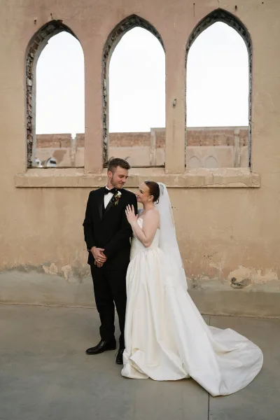 Couple portrait as the bride and groom pose, her long veil trailing as they stand beside arched windows set in a stucco wall.