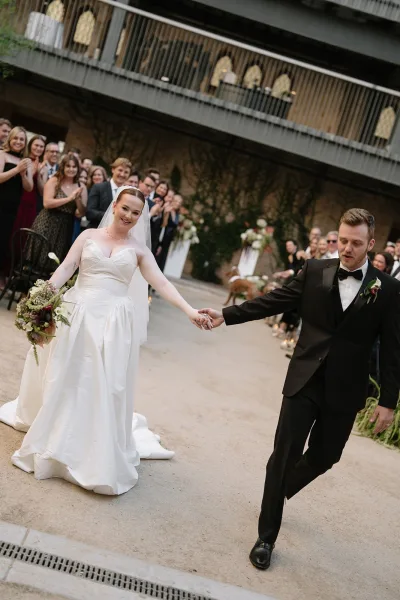 Wedding recessional during a just married exit, newlyweds walk the aisle with their dog against an ivy wall in an outdoor courtyard.