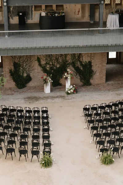 Ceremony setup in industrial ceremony decor with black chairs flanking a wide aisle before a brick wall and metal balcony backdrop.