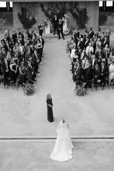 Ceremony processional as the bride walking down aisle with a flowing veil, groom waits at the altar before a brick wall draped in climbing vines.