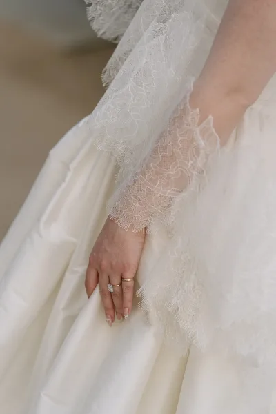 Wedding dress detail featuring a lace bridal sleeve and square engagement ring over an ivory gown with floral lace edging.