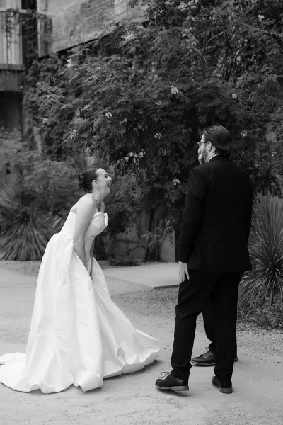 Couple portrait capturing a candid wedding moment as the bride laughs in strapless bridal gown while the groom with glasses smiles by garden greenery.