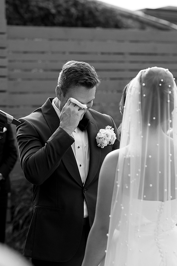 First look moment as the groom wiping tears with a handkerchief in tuxedo and bow tie, bride from behind in pearl veil on patio