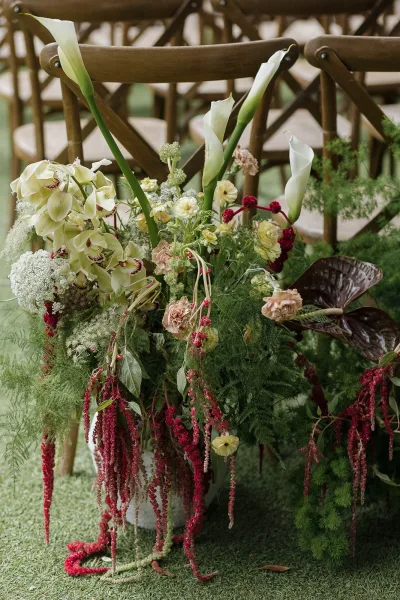 Ceremony aisle flowers form an elegant aisle marker arrangement with white calla lilies beside wood crossback chairs on a grass lawn.