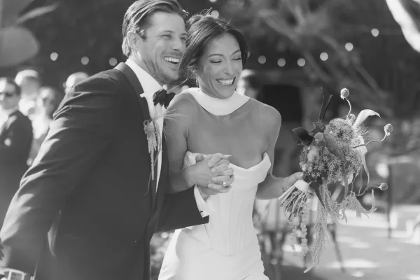 Wedding recessional ceremony exit moment as bride and groom walk the aisle holding hands, bride wearing a bridal choker amid trees and string lights.