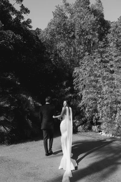 Couple portrait of the bride and groom walking hand in hand along a garden pathway, her long veil trailing as trees frame their quiet stroll.