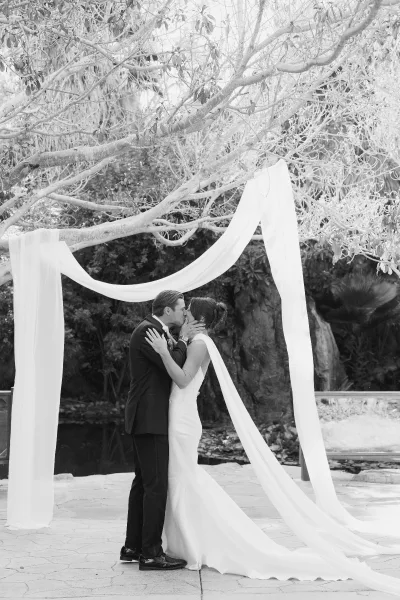 Wedding kiss portrait of bride and groom sharing their ceremony first kiss beneath a draped fabric arch on a stone patio by a garden pond.