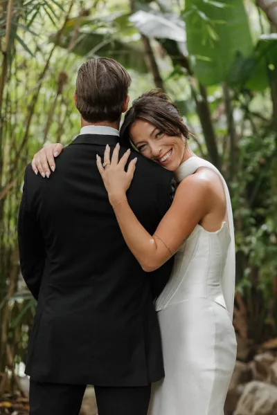 Couple portrait of a bride hugging groom, her sleek satin wedding dress shining, framed by tropical garden bamboo and lush foliage.