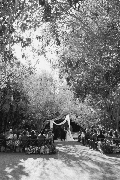 Ceremony moment at an outdoor wedding ceremony as the couple exchange vows beneath a draped fabric arch in a sunlit garden under trees.