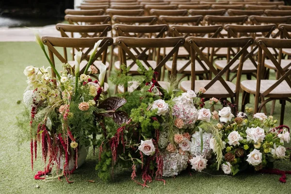 Ceremony aisle flowers shape floral aisle arrangements flanking wooden crossback chairs, gathered in low, grounded clusters with organic lines.