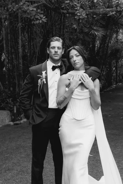 Couple portrait of a bride and groom in a garden, standing arm over shoulder on a stone pathway, her shoulder cape draping elegantly.