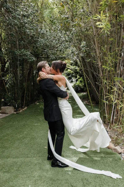 Wedding kiss portrait as the groom lifts the bride, her long veil trailing on the grass, framed by a serene bamboo grove.