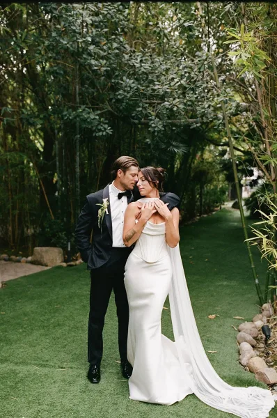 Couple portrait of a bride and groom embrace, her bridal cape veil flowing as they stand on a garden lawn path bordered by bamboo greenery.