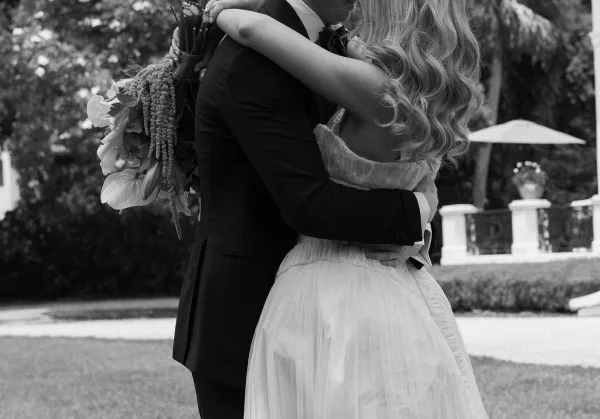 Wedding hug as bride and groom embrace from behind, her bouquet with greenery draped over a tulle gown in a garden setting
