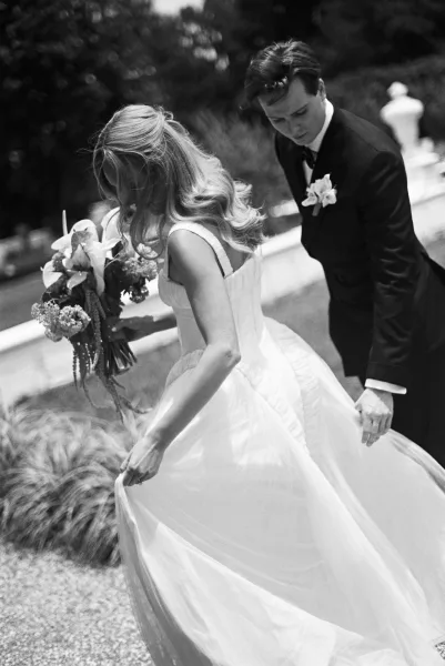 Couple portrait in a black and white wedding portrait, bride holding bouquet while lifting her gown beside groom in tux on stone garden steps