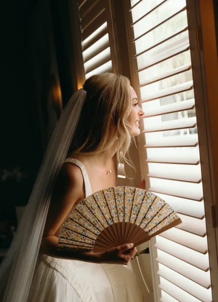Bridal portrait of a bride by window in natural light, side profile with long veil and hand fan against white window shutters
