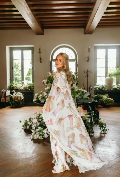 Bridal portrait of a bride in a printed robe with long wavy hair, standing by a grand piano amid floral arrangements and greenery garlands