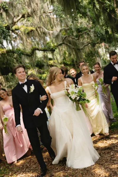 Wedding recessional as bride and groom walk the aisle under mossy trees, bride in veil holding white calla lily bouquet, party behind