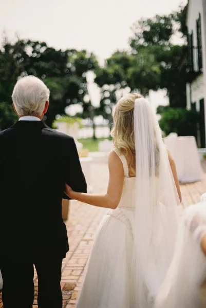 Wedding processional with bride walking down aisle on her father’s arm, chapel-length veil and tuxedo on a brick walkway by trees