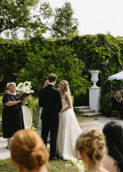 Wedding vows as bride in veil and groom in black tux face each other while officiant reads before white florals in a garden setting