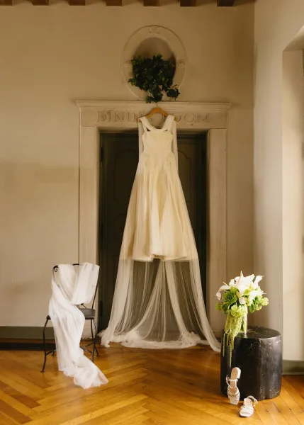 Wedding dress hanging on a wooden hanger with long veil, bouquet in vase, and bridal shoes in an arched doorway setting